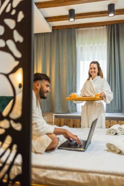 Kız arkadaşı ona kahvaltı hazırlarken, beyaz bornozlu genç adam yatakta gülümsüyor..