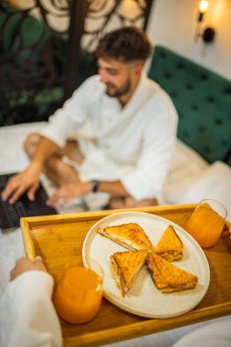 Kız arkadaşı ona kahvaltı hazırlarken, beyaz bornozlu genç adam yatakta gülümsüyor..