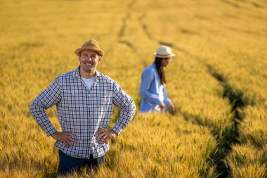 Üst düzey bir adam altın buğday tarlasında dikilirken kameraya güvenle gülümsüyor. Arka planda özenle çalışan Taylandlı bir kadın çiftçi ile birlikte..