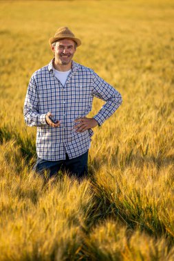 Üst düzey bir adam altın buğday tarlasında dikilirken kameraya güvenle gülümsüyor. Arka planda özenle çalışan Taylandlı bir kadın çiftçi ile birlikte..