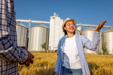 İki işçi buğday kaplı bir alanda büyük siloların önünde duruyor ve gelecek görevleri aktif olarak tartışıyorlar..