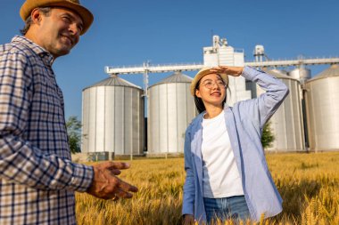 İki işçi buğday kaplı bir alanda büyük siloların önünde duruyor ve gelecek görevleri aktif olarak tartışıyorlar..