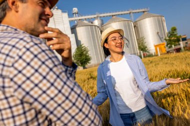 İki işçi buğday kaplı bir alanda büyük siloların önünde duruyor ve gelecek görevleri aktif olarak tartışıyorlar..