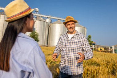 İki işçi buğday kaplı bir alanda büyük siloların önünde duruyor ve gelecek görevleri aktif olarak tartışıyorlar..