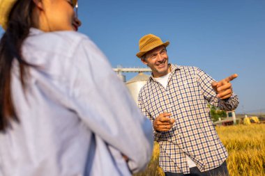 İki işçi buğday kaplı bir alanda büyük siloların önünde duruyor ve gelecek görevleri aktif olarak tartışıyorlar..