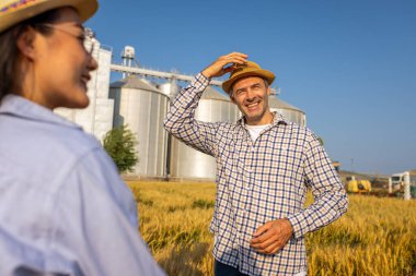 İki işçi buğday kaplı bir alanda büyük siloların önünde duruyor ve gelecek görevleri aktif olarak tartışıyorlar..