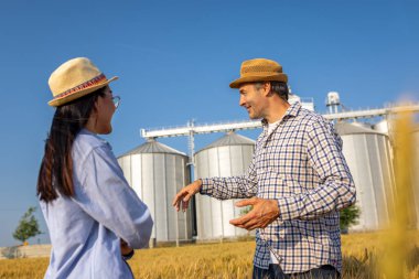 İki işçi buğday kaplı bir alanda büyük siloların önünde duruyor ve gelecek görevleri aktif olarak tartışıyorlar..