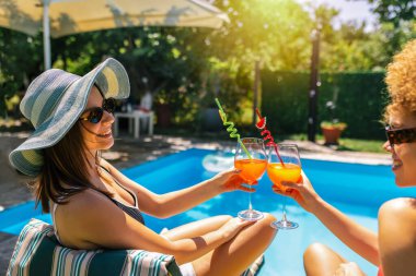 Mayolu iki kız arkadaş havuz kenarında eğleniyor, renkli kokteyller içiyor ve güneşli, neşeli bir yaz anının tadını çıkarıyor..