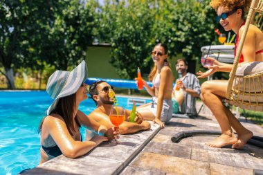 Bir grup genç havuz kenarında takılıyor, bazıları suda bacakları olan, renkli kokteyllerin keyfini çıkaran ve güneşli bir günde birlikte dinlenen insanlar..