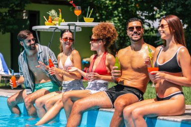 Mayolu bir grup genç, havuz kenarında bacakları suda, renkli kokteyllerin tadını çıkarıyor ve güneşli bir günde birlikte eğleniyor..