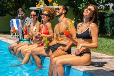 Mayolu bir grup genç, havuz kenarında bacakları suda, renkli kokteyllerin tadını çıkarıyor ve güneşli bir günde birlikte eğleniyor..