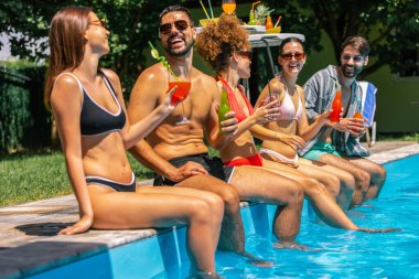 Mayolu bir grup genç, havuz kenarında bacakları suda, renkli kokteyllerin tadını çıkarıyor ve güneşli bir günde birlikte eğleniyor..