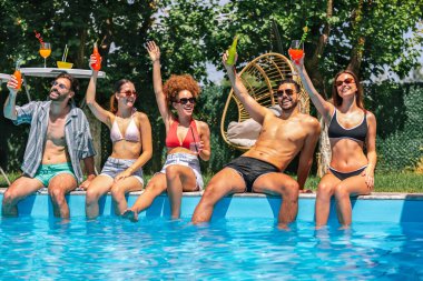 Mayolu bir grup genç, havuz kenarında bacakları suda, renkli kokteyllerin tadını çıkarıyor ve güneşli bir günde birlikte eğleniyor..
