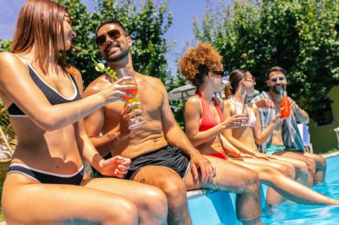 Mayolu bir grup genç, havuz kenarında bacakları suda, renkli kokteyllerin tadını çıkarıyor ve güneşli bir günde birlikte eğleniyor..