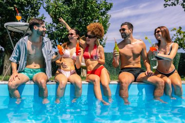 Mayolu bir grup genç, havuz kenarında bacakları suda, renkli kokteyllerin tadını çıkarıyor ve güneşli bir günde birlikte eğleniyor..