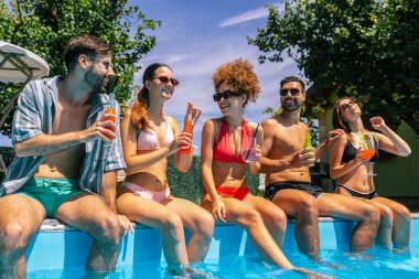 Mayolu bir grup genç, havuz kenarında bacakları suda, renkli kokteyllerin tadını çıkarıyor ve güneşli bir günde birlikte eğleniyor..