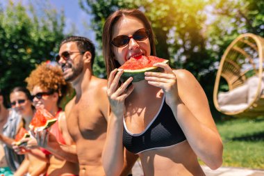 Mayolu bir grup genç havuz kenarında taze karpuzun tadını çıkarıyor güneşli bir günde eğlenceli ve ferahlatıcı bir yaz anını paylaşıyorlar..