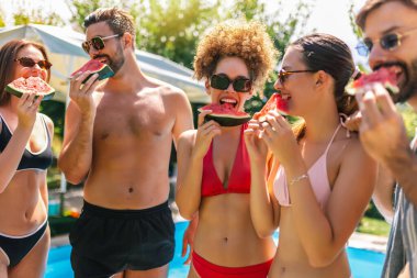 Mayolu bir grup genç havuz kenarında taze karpuzun tadını çıkarıyor güneşli bir günde eğlenceli ve ferahlatıcı bir yaz anını paylaşıyorlar..