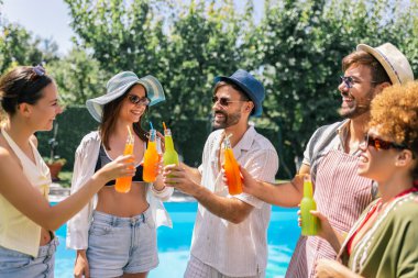 Bir grup genç havuz kenarında eğleniyor, mayo, şapka ve güneş gözlüğü takıyor ve pipetle renkli kokteyller içiyor..