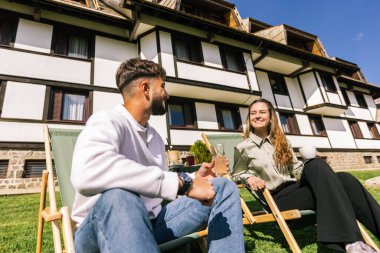 Genç bir adam ve bir kadın, hafta sonu kaçamağı sırasında şezlonglarda dinleniyorlar. Doğa ve temiz havayla çevrili olarak sıcak kahvenin ve ferahlatıcı meyve suyunun tadını çıkarırlar..