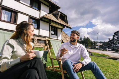 Genç bir adam ve bir kadın, hafta sonu kaçamağı sırasında şezlonglarda dinleniyorlar. Doğa ve temiz havayla çevrili olarak sıcak kahvenin ve ferahlatıcı meyve suyunun tadını çıkarırlar..