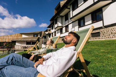Genç bir adam ve kadın güneşli bir hafta sonu kaçışı sırasında bir dağ otelindeki şezlonglarda dinleniyorlar..