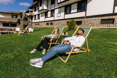 Genç bir adam ve kadın güneşli bir hafta sonu kaçışı sırasında bir dağ otelindeki şezlonglarda dinleniyorlar..