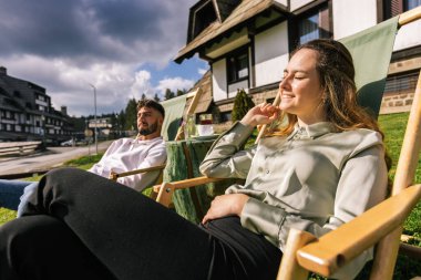 Genç bir adam ve kadın güneşli bir hafta sonu kaçışı sırasında bir dağ otelindeki şezlonglarda dinleniyorlar..