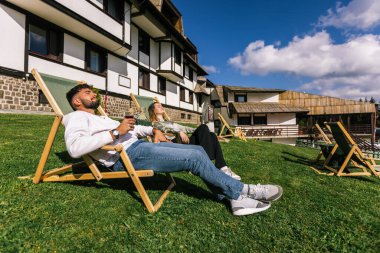Genç bir adam ve kadın güneşli bir hafta sonu kaçışı sırasında bir dağ otelindeki şezlonglarda dinleniyorlar..