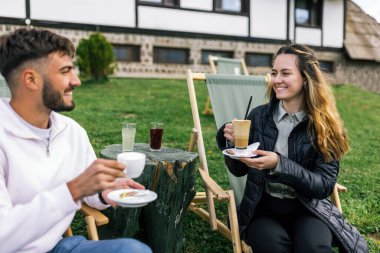 Genç bir adam ve bir kadın, hafta sonu kaçamağı sırasında şezlonglarda dinleniyorlar. Doğa ve temiz havayla çevrili olarak sıcak kahvenin ve ferahlatıcı meyve suyunun tadını çıkarırlar..