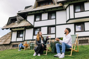 Genç bir adam ve bir kadın, hafta sonu kaçamağı sırasında şezlonglarda dinleniyorlar. Doğa ve temiz havayla çevrili olarak sıcak kahvenin ve ferahlatıcı meyve suyunun tadını çıkarırlar..