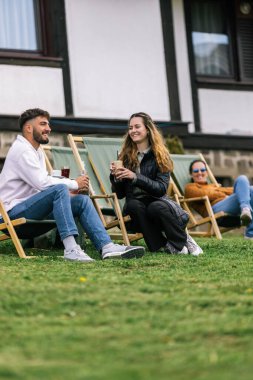 Genç bir adam ve bir kadın, hafta sonu kaçamağı sırasında şezlonglarda dinleniyorlar. Doğa ve temiz havayla çevrili olarak sıcak kahvenin ve ferahlatıcı meyve suyunun tadını çıkarırlar..