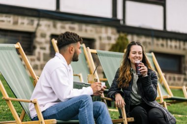 Genç bir adam ve bir kadın, hafta sonu kaçamağı sırasında şezlonglarda dinleniyorlar. Doğa ve temiz havayla çevrili olarak sıcak kahvenin ve ferahlatıcı meyve suyunun tadını çıkarırlar..