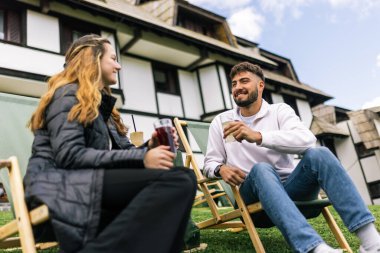 Genç bir adam ve bir kadın, hafta sonu kaçamağı sırasında şezlonglarda dinleniyorlar. Doğa ve temiz havayla çevrili olarak sıcak kahvenin ve ferahlatıcı meyve suyunun tadını çıkarırlar..