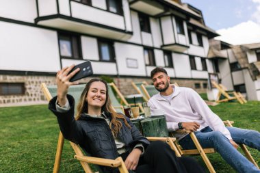 Genç bir erkek ve kadın manzaralı bir dağ evinde hafta sonu kaçamağının tadını çıkarıyorlar. Şezlonglarda uzanırken, o anı, akıllı telefonlarıyla neşeli bir selfie çekerek yakalıyorlar..