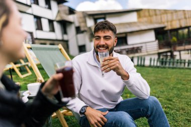 Genç bir adam ve bir kadın, hafta sonu kaçamağı sırasında şezlonglarda dinleniyorlar. Doğa ve temiz havayla çevrili olarak sıcak kahvenin ve ferahlatıcı meyve suyunun tadını çıkarırlar..