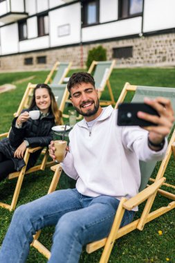 Genç bir erkek ve kadın manzaralı bir dağ evinde hafta sonu kaçamağının tadını çıkarıyorlar. Şezlonglarda uzanırken, o anı, akıllı telefonlarıyla neşeli bir selfie çekerek yakalıyorlar..