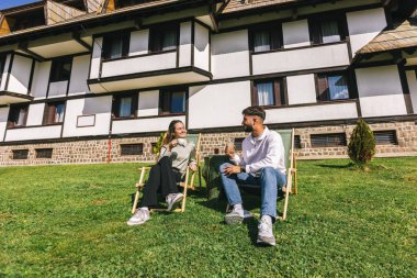 Genç bir adam ve bir kadın, hafta sonu kaçamağı sırasında şezlonglarda dinleniyorlar. Doğa ve temiz havayla çevrili olarak sıcak kahvenin ve ferahlatıcı meyve suyunun tadını çıkarırlar..