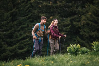 Sırt çantaları, yürüyüş direkleri ve açık hava kıyafetleriyle donatılmış genç bir erkek ve kadın, bereketli doğayla çevrili bir dağ yolu boyunca birlikte yürüyorlar. Çevrelerindeki manzaranın güzelliğine hayran olmak için dururlar..