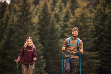 Sırt çantaları, yürüyüş direkleri ve açık hava kıyafetleriyle donatılmış genç bir erkek ve kadın, yemyeşil doğayla çevrili bir dağ yolunda birlikte yürüyorlar.. 