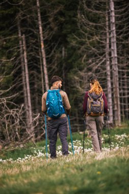 Genç bir adam ve kadın, sırt çantaları, yürüyüş direkleri ve açık hava kıyafetleriyle dolu manzaralı bir dağ yolunda birlikte yürüyüş yapıyorlar. Fotoğraf arkadan çekilmiş..