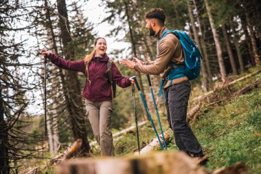 Genç bir adam, ormanlık bir dağ patikasının zorlu bir bölümünde bir kadına yardım ediyor. İkisi de tam teçhizatlı yürüyüş kıyafetleri içinde, sırt çantaları, yürüyüş kıyafetleri ve direkler..