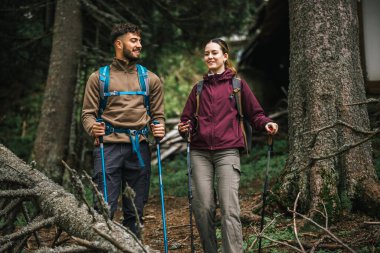 Sırt çantaları, yürüyüş direkleri ve açık hava kıyafetleriyle donatılmış genç bir erkek ve kadın, yemyeşil doğayla çevrili bir dağ yolunda birlikte yürüyorlar.. 