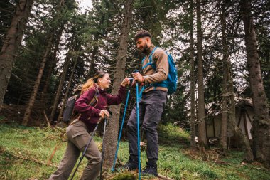 Genç bir adam, ormanlık bir dağ patikasının zorlu bir bölümünde bir kadına yardım ediyor. İkisi de tam teçhizatlı yürüyüş kıyafetleri içinde, sırt çantaları, yürüyüş kıyafetleri ve direkler..