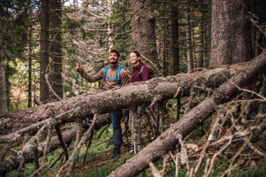 Sırt çantaları, yürüyüş direkleri ve açık hava kıyafetleriyle donatılmış genç bir erkek ve kadın, yemyeşil doğayla çevrili bir dağ yolunda birlikte yürüyorlar.. 
