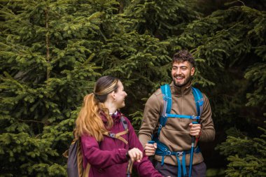 Sırt çantaları, yürüyüş direkleri ve yürüyüş elbiseleriyle donatılmış genç bir adam ve kadın yoğun çalılarla çevrili engebeli bir dağ yolunda dikkatle ilerliyorlar.. 