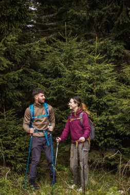 Sırt çantaları, yürüyüş direkleri ve yürüyüş elbiseleriyle donatılmış genç bir adam ve kadın yoğun çalılarla çevrili engebeli bir dağ yolunda dikkatle ilerliyorlar.. 