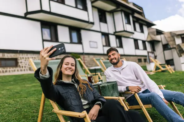 Genç bir erkek ve kadın manzaralı bir dağ evinde hafta sonu kaçamağının tadını çıkarıyorlar. Şezlonglarda uzanırken, o anı, akıllı telefonlarıyla neşeli bir selfie çekerek yakalıyorlar..