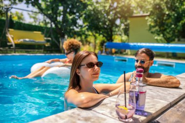 Gençler havuzda güneşli bir yaz gününün tadını çıkarıyorlar. Bir kız ve erkek arkadaşının yüzen ve rahatlatan arkadaşlarıyla birlikte mor kokteyller içip gülmelerine odaklanıyorlar..