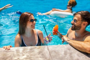 Gençler havuzda güneşli bir yaz gününün tadını çıkarıyorlar. Bir kız ve erkek arkadaşının yüzen ve rahatlatan arkadaşlarıyla birlikte mor ve turuncu kokteyller içip gülmelerine odaklanıyorlar..